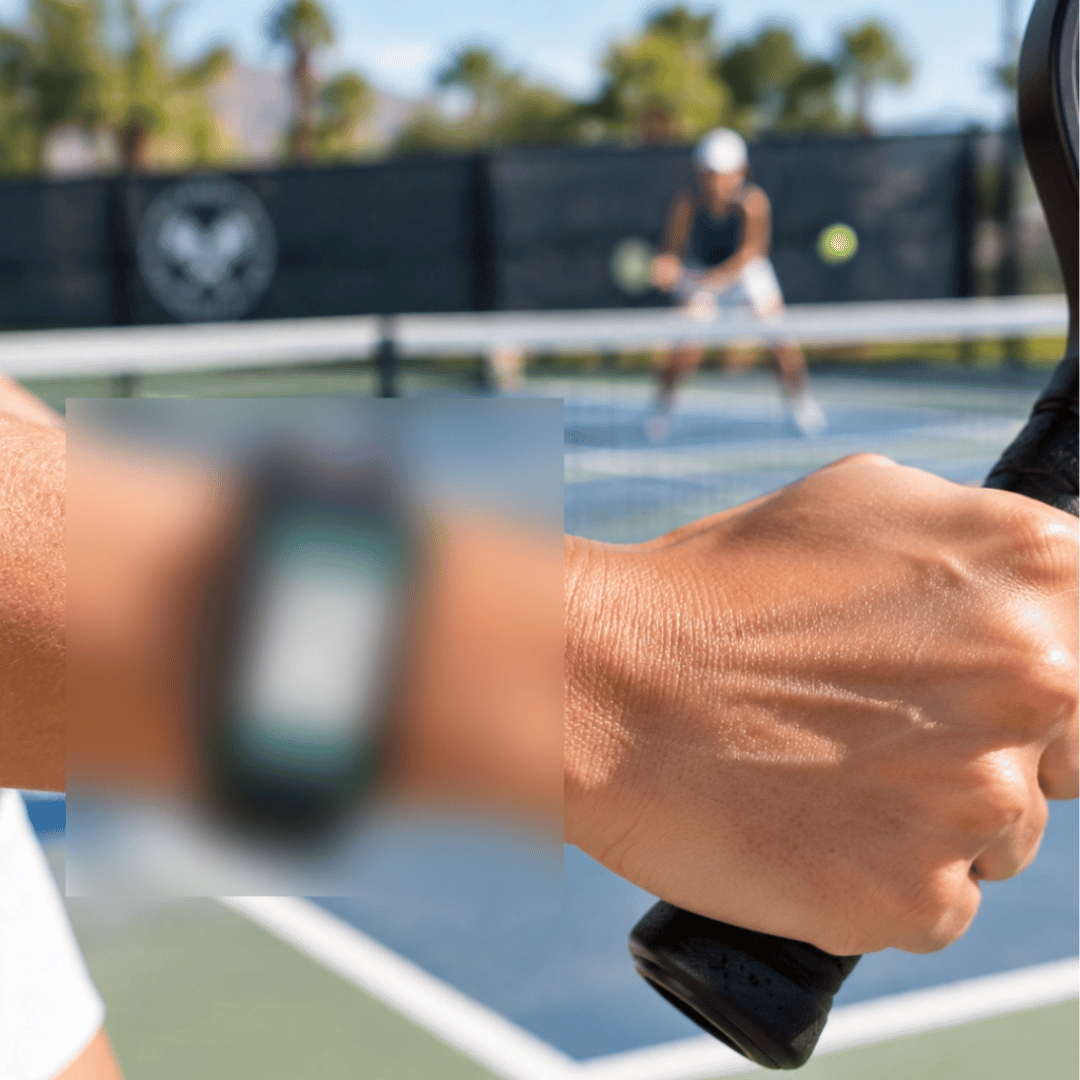 A close-up of a smartwatch worn on a wrist, with a blurred background showing a pickleball court and a player holding a pickleball paddle. The watch has a dark green and black face with a white screen area.